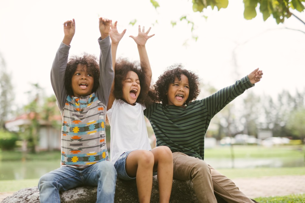 three children of color are sitting side by side, arms up in a joyful pose